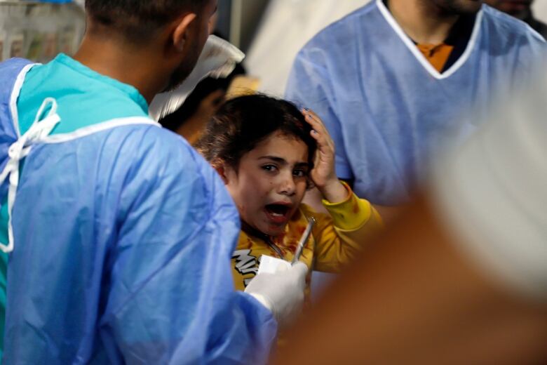 A young Palestinian girl with blood on her clothes cries and holds her head as two people in medical gowns work on treating her wounds.