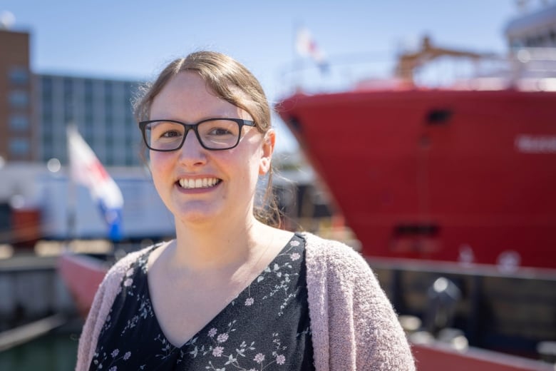 A woman wearing dark-framed glasses, a floral short and a pink cardigan smiles. Behind her, a large red ship can be seen.