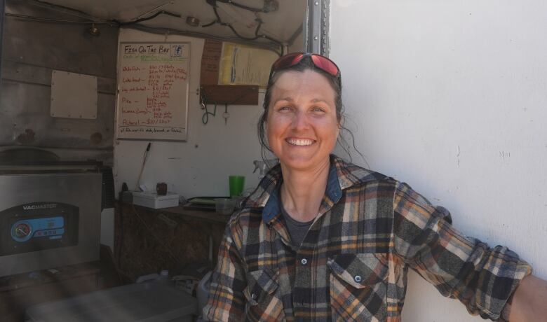 A woman in plaid with sunglasses on her head smiles in to the camera. She's standing in the doorway of a trailer, in the background you can see equipment and a white board with fish prices.