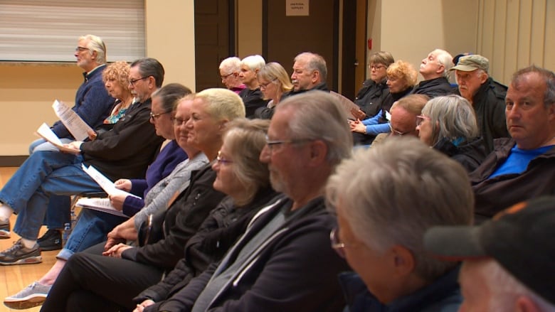 A side-view of a group of people seated. The are listening to and watching a meeting.