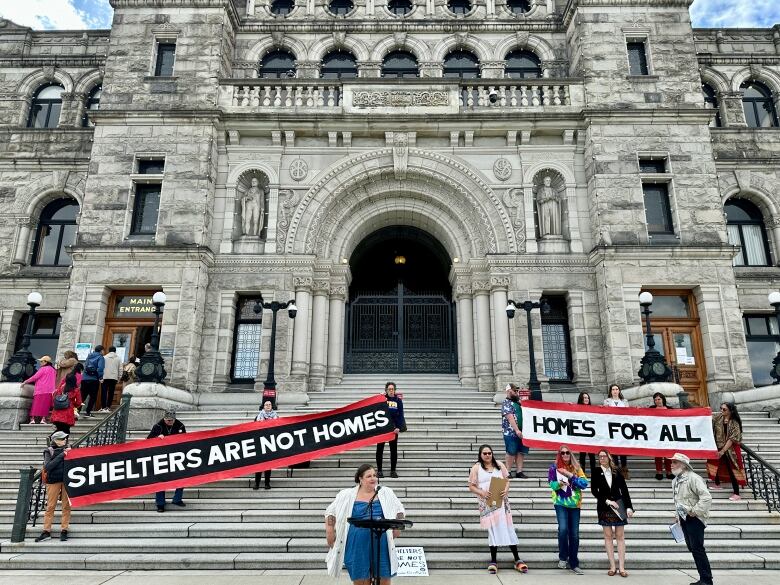 A group of people stand on the steps of a large, ornate stone building. They hold two banners, reading 