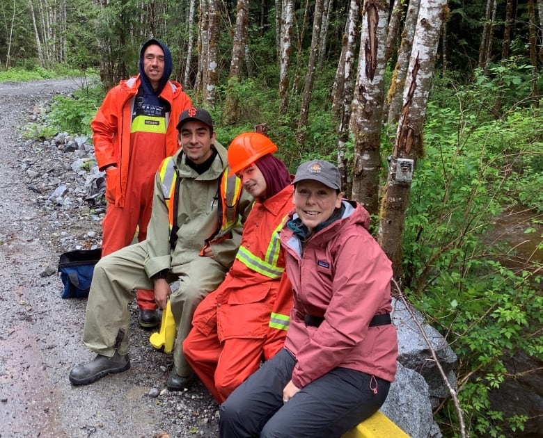 Four people wearing rain jackets and high-visibility gear pose for a photo on a road through a forest.