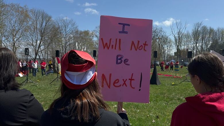 A person with brown hair stands outside with their back to the camera, holding up a sign that says I will not be next.