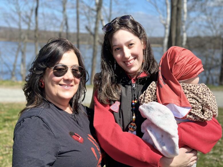 A young woman with long brown hair stands outside in the sun holding a baby with a red scarf around its head. An older woman with short brown hair stands smiling next to them.