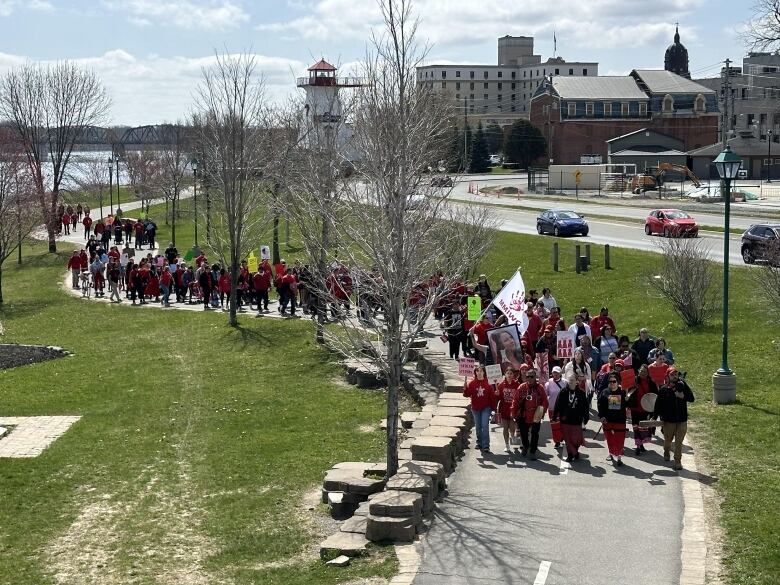 A crowd of people wearing red and carrying flags and signs marches down a winding, paved path on a sunny day.