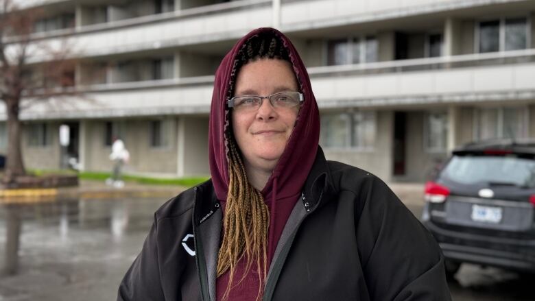 A woman in a hood stands in front of an apartment.