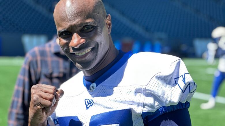 A bald man in a football uniform smiles and makes fist with his right hand as he poses for a photo.