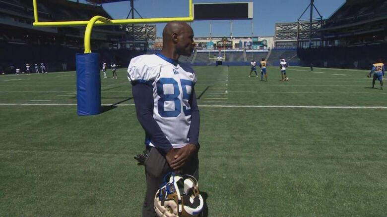 A football player holds his helmet at his waist while standing in the end zone and looking back toward the field.