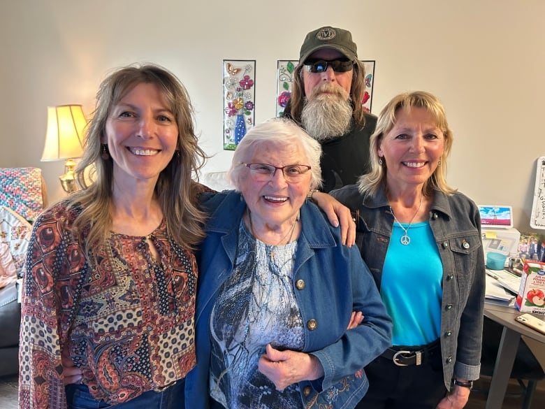 Woman, 86, with grey hair and glasses stands surrounded by her three children. The woman on the left has blond hair, the man in the back has a long beard, baseball cap and glasses, The woman on the right is blonde. 