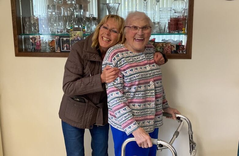 Woman on the left with blond hair, brown jacket and jeans stands hugging a woman with grey hair, colourful sweater and blue pants. The woman on the right is standing with the help of a walker. 