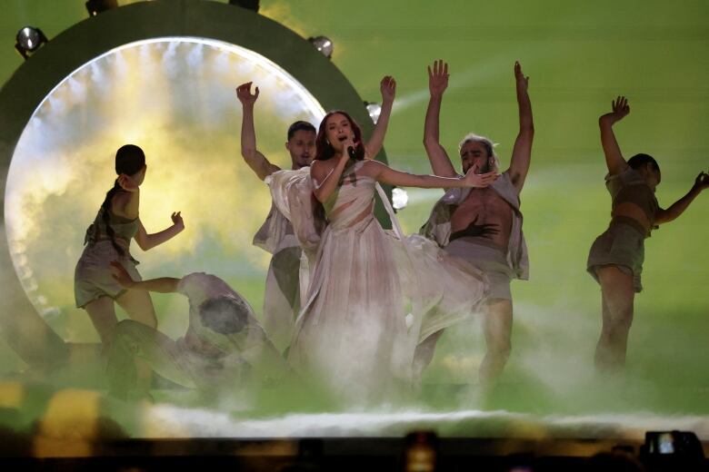 A woman in a white dress sings on stage, flanked by dancers.