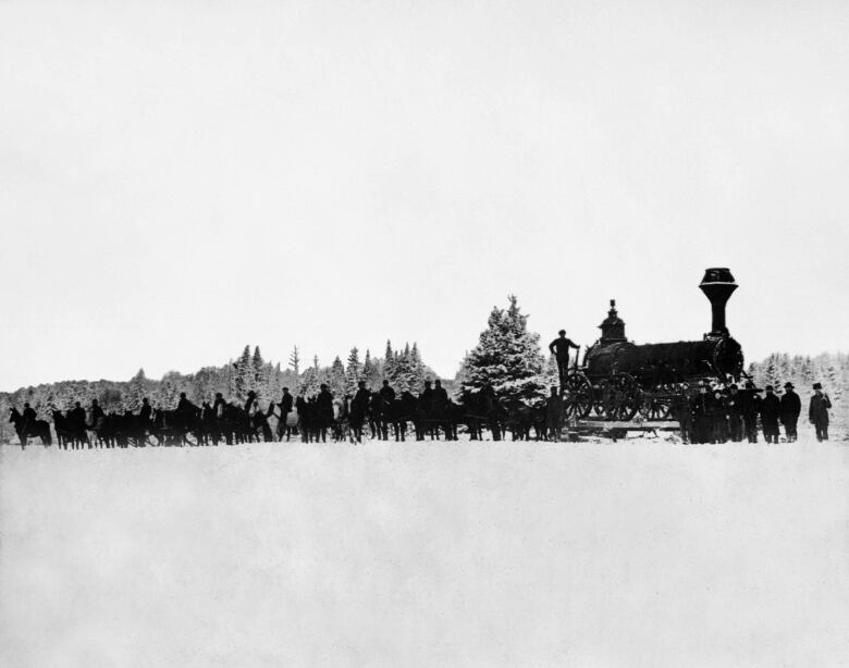 A black and white sideview photo of a procession of a few dozen people and some horses to the left and a train engine at right. The foreground is a snowy open field. The background is a line of snowcovered evergreens against a light gray sky.