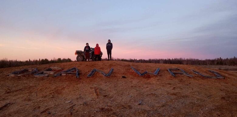 Three people are seen standing on a hill. A message is spelled out with pieces of wood that says 