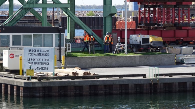 Four people in high-visibility vests and hard hats next to a large, green, metal bridge.