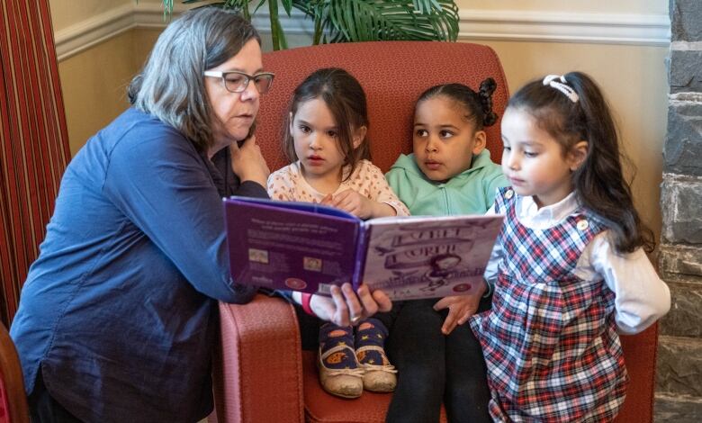 Kids sit in a chair and look at a book together with their teacher.