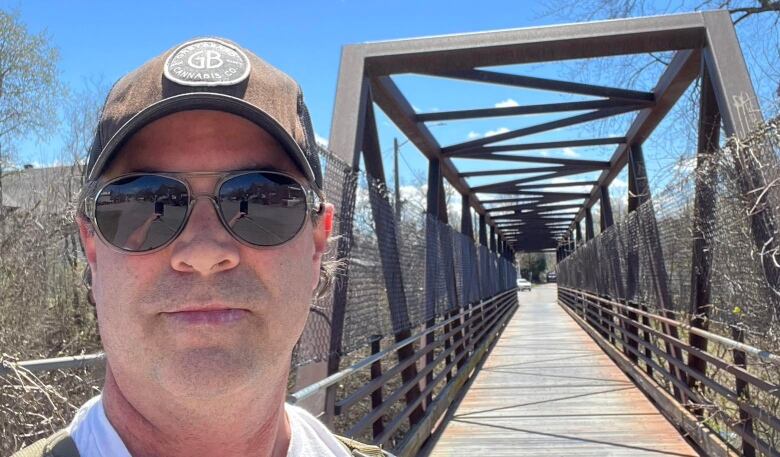 A man wearing a hat and sunglasses stands next to a bridge