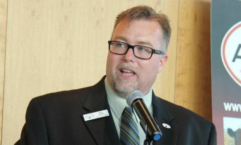 A man stands behind a podium and a microphone, wearing a suit.