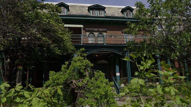 A red brick building with a green veranda and second storey stands behind some trees and shrubs