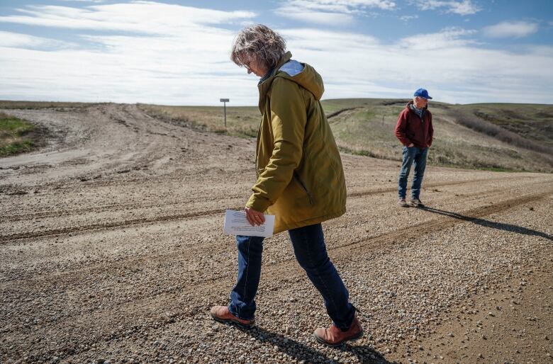 A woman and a man walk across a gravel country road.