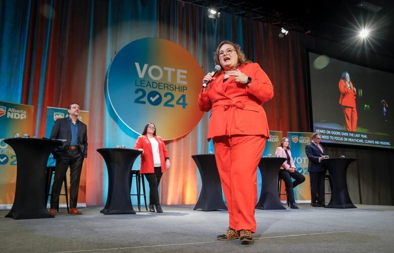 a woman speaks into a microphone while others await their turn at a debate.