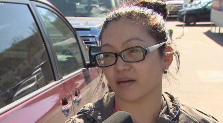 A woman with olive skin and dark brown hair, tied in a bun, is wearing glasses and a black jacket. She is standing in parking lot, under the sunshine, beside her red minivan.