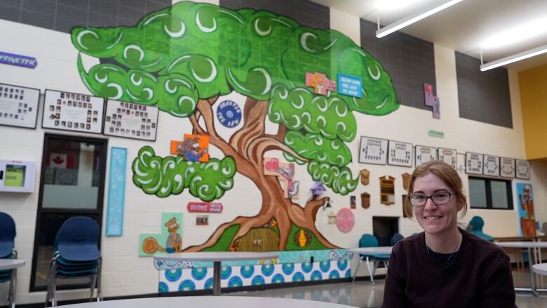 A woman sitting down in a crew neck sweater in front of a large mural of a tree. 