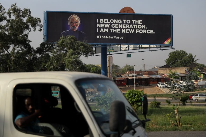 Cars on the streets of Accra, Ghana, drive past a billboard showing a figure wearing a traditional face mask with the words 