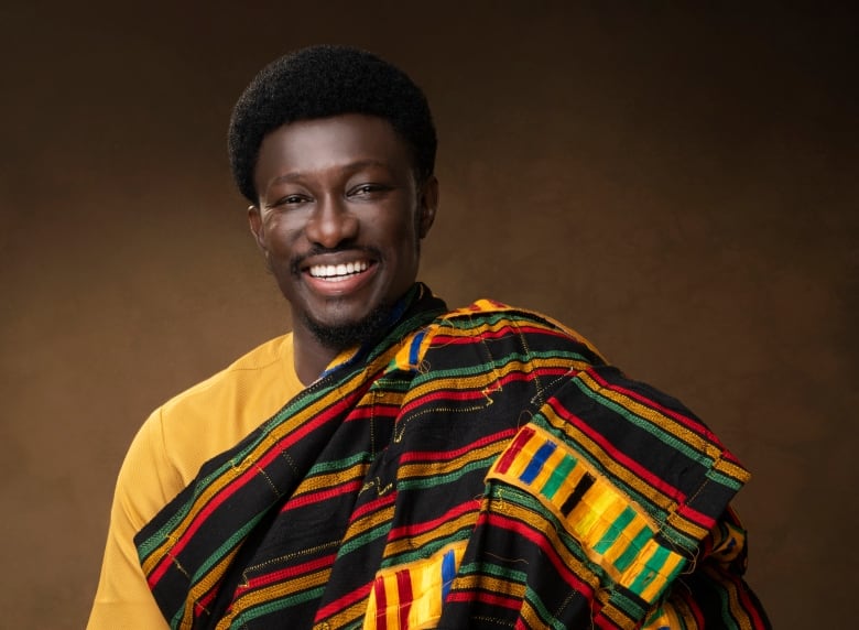 An Ghanian man wearing a yellow t-shirt with a traditional Kente cloth robe smiles. 