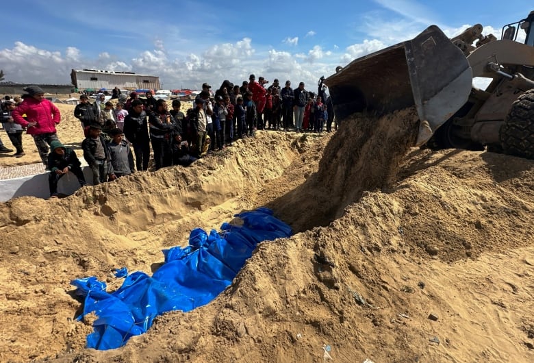 A bulldozer piles dirt on top of a row of blue body bags in a mass grave as people stand alongside watching. 