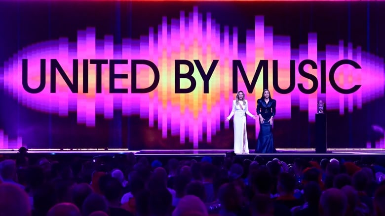 Two women in gowns stand on a stage in front of a giant projection of the Eurovision slogan, 