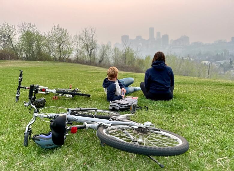 A couple sits on the green grass, a man with red hair and a women with brown hair, looking up at the skyline, that's smoky their bikes on the grass.