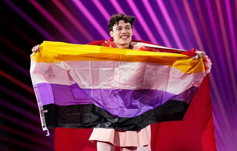 A smiling person holds up a flag made up of horizontal yellow, white purple and black stripes representing the non-binary community.  