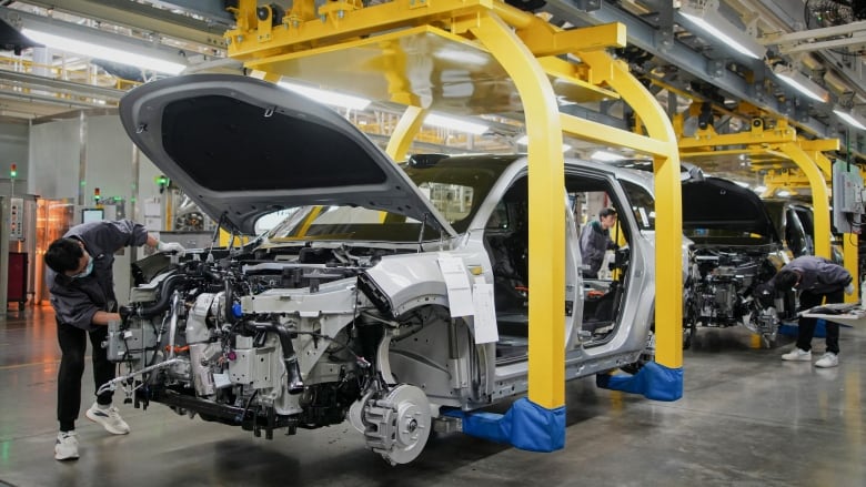 A worker is shown examining the inner workings of the front of an automobile in a factory setting.