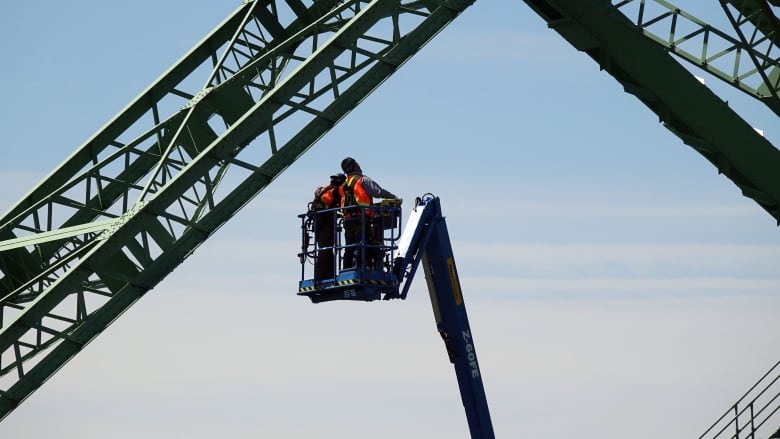 Two people in bright orange vests in an aerial lift, examining part of a large, green, metal bridge.