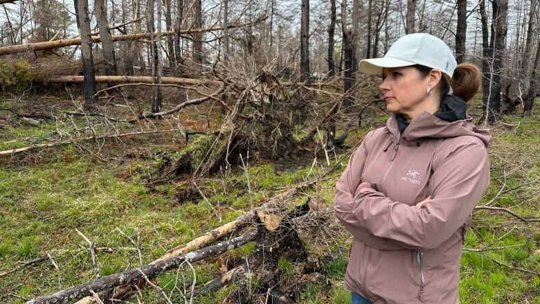 A woman is shown standing in a grassy area alongside charred trees in the background and others that fell down during the wildfire.