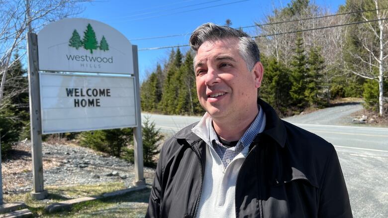 A man is shown by a community welcome sign by the road which says 