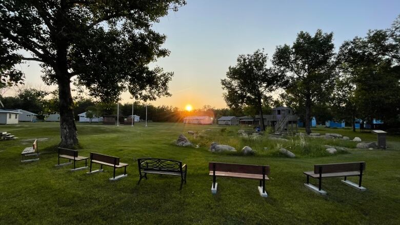 A sun sets over a grassy field with benches and trees.