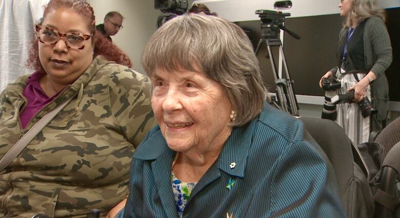 An older woman smiles into the camera, with media personnel in the background.