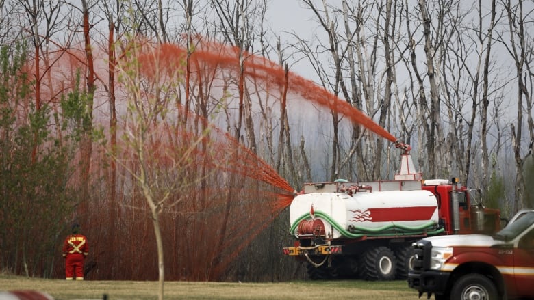 A pumper truck sprays bright red fire retardant on trees. The sky is smoky.