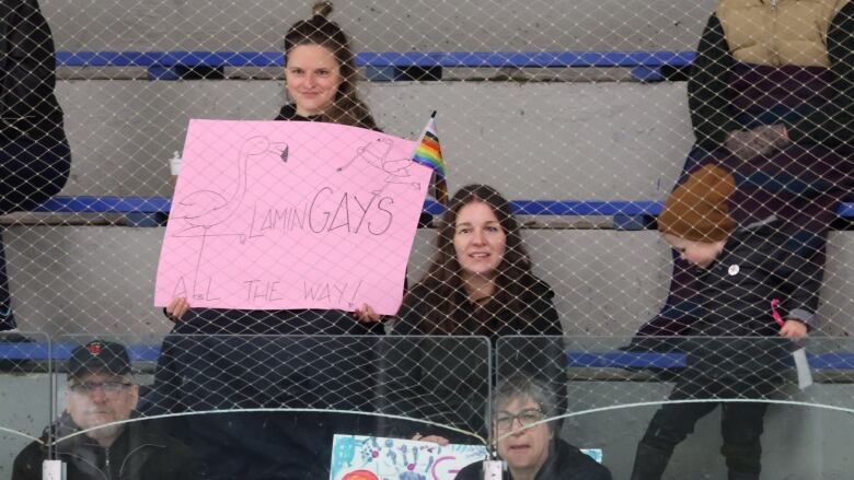 A person sits in the hockey stands holding a pride flag and a pink sign that says 