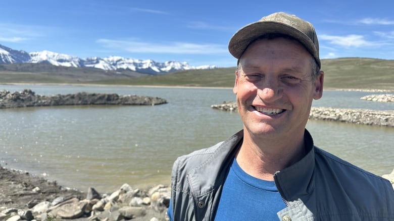 A man wearing a ballcap stands in front of a small lake with mountains in the background.