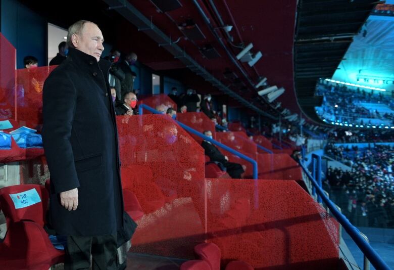 A man in a dark coat stands along near a red backdrop looking down over a stadium.