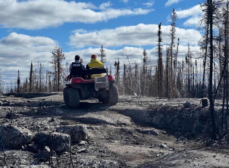 A police officer and a search and rescue member seen from behind driving a quad through burnt forest.