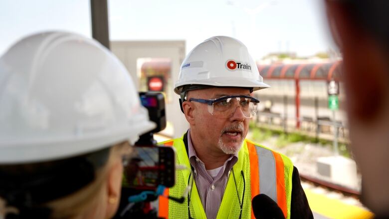 A man in a white hard hat and florescent vest speaks to reporters