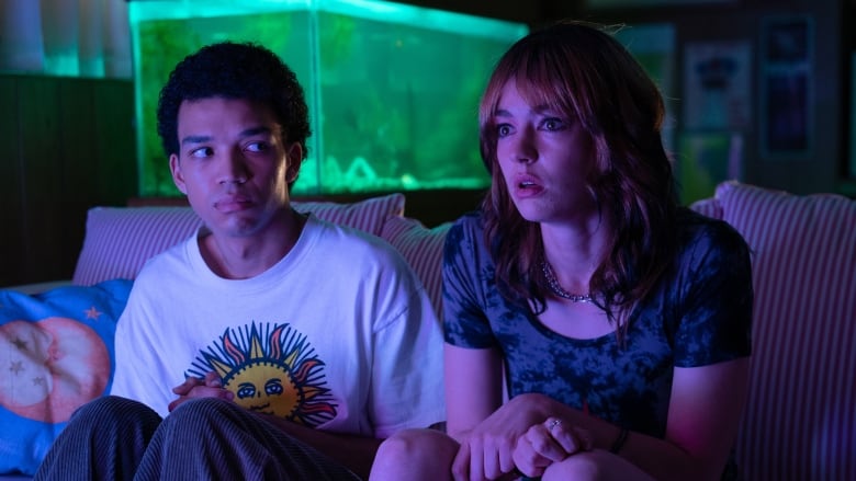 Two teenagers sit in a dark room. Behind them is a fish tank lit up green. 