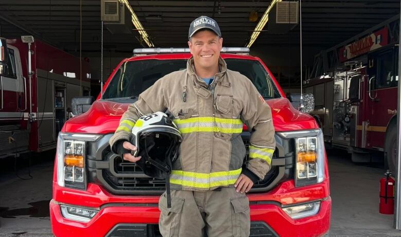 A man in firefighting gear stands before a truck, helmet tucked under his right arm.