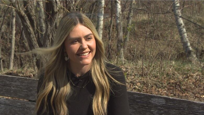 A woman with long blonde hair smiles while sitting on a park bench.