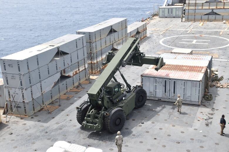 Shot from above, three men including two in military uniform on a large platform with a helicopter landing circle visible. A military vehicle with a lifting mechanism and several large containers are also visible.