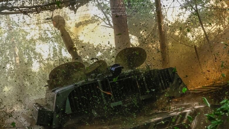 A tank sits in heavy brush. Foliage and dirt fill the air.