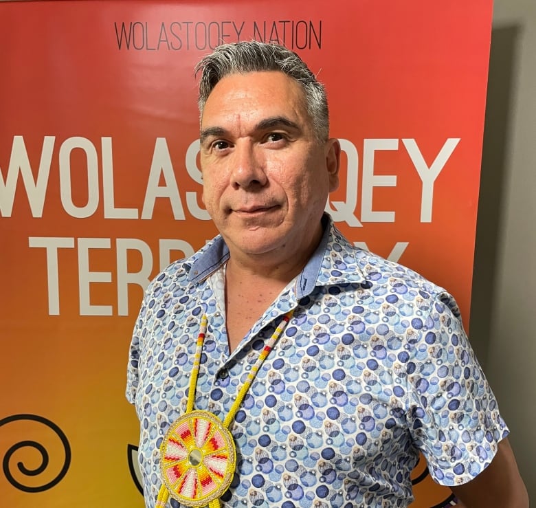 A man with short, grey hair and wearing a blue shirt with a yellow, beaded necklace stands in front of an orange poster.